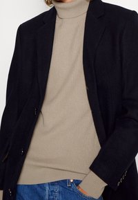 Schwarzer Blazer mit reißverschlussartigen Revers über einem beigen gerippten Rollkragenpullover, kombiniert mit blauen Jeans. Texturierte Stoffe sind in beiden Schichten sichtbar.