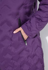 Chaqueta de plumas púrpura con un diseño acolchado, bolsillo lateral con cremallera y detalles en cremallera negra en la parte delantera y en los bolsillos.
