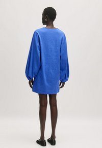 Blaues übergroßes Kleid aus strukturiertem Stoff, mit langen Puffärmeln und einem runden Ausschnitt. Schwarze Schuhe ergänzen die Schlichtheit des Outfits.