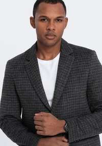 Grau karierter Blazer aus strukturiertem Stoff, mit einem Reverskragen und zwei Knöpfen. Wird über einem weißen T-Shirt getragen, das einen modernen Schnitt zeigt.