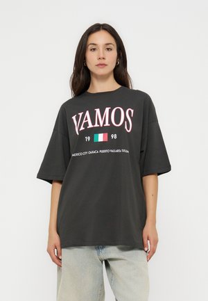 Frau trägt ein schwarzes oversized T-Shirt mit dem Schriftzug „VAMOS“, mexikanischer Flagge, „1998“ und Stadtnamen auf der Vorderseite, kombiniert mit hellen Jeans.