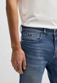 Blå denimjeans med en lätt blekning, med fem fickor, knappgylf och en liten svart etikett på midjebandet. Modellen har handen synlig.