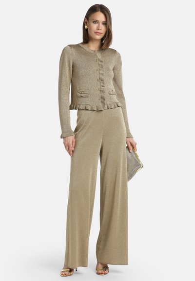 Auksinė megzta kardiganas su pūstomis apykaklėmis ir priekiniais mygtukais, derinamas su aukštu juosmeniu plačiomis kelnėmis iš atitinkamos auksinės medžiagos.