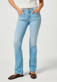 Jean en denim bleu clair évasé avec une taille mi-haute, cinq poches et une fermeture à bouton à l'avant. Porté avec des sandales plates tressées brunes.