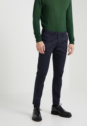 Mann in dunkelblauen Hosen, schwarzen Leder-Schnürschuhen und grünem Langarmpullover, der vor einem weißen Hintergrund steht.