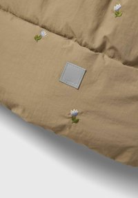 Tissu matelassé beige avec des broderies florales en bleu et blanc, portant une étiquette rectangulaire grise cousue dans le design.