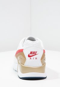 Baskets blanches avec des accents dorés et rouges, présentant une tige texturée, un col rembourré et une semelle en caoutchouc. Logo Nike en rouge à l'arrière.