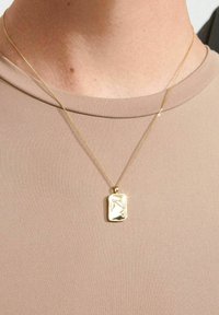 Goldene Halskette mit einem rechteckigen Anhänger, der eine strukturierte Oberfläche und ein graviertes Design aufweist. Die Kette ist zart und flach. Getragen über einem beigen Oberteil.