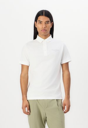Polo bianco a maniche corte, colletto classico e piccolo logo ricamato sul petto, abbinato a pantaloni verde chiaro.