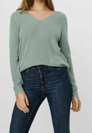 Vrouw gekleed in een lichtgroene geribbelde gebreide V-hals trui en donkerblauwe spijkerbroek, staand voor een effen achtergrond.