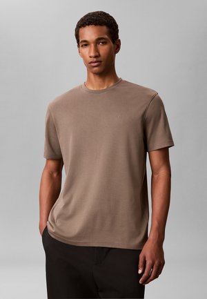 Giovane uomo con capelli corti e ricci che indossa una maglietta marrone tinta unita a maniche corte e pantaloni neri, in piedi davanti a uno sfondo grigio neutro.