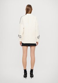 Pleteni kardigan v kremni barvi z teksturiranimi vzorci, oversize kroj, kontrastni črni detajli na rokavih ter rebrastimi manšeti.