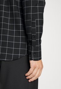 Camicia a scacchi nera con motivo a griglia bianca; presenta polsini con bottoni e tessuto liscio; l'orlo curva nella parte inferiore.