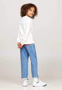 Felpa bianca con dettagli rossi, jeans larghi blu chiaro e sneakers bianche con accenti navy. Capelli ricci incorniciano il viso.