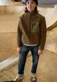 Bruine fleece jack met groene accenten, ritszakken; blauwe spijkerbroek en witte sneakers. Binnen skatepark met gladde oppervlakken.