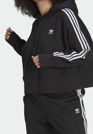 Person iført sort Adidas-hættetrøje og bukser med hvide tre-stribede detaljer på ærmer og ben, vist fra nakke til talje.