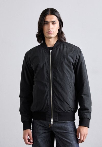 Černá bomber bunda se zapínáním na zip, žebrovanými manžetami a texturovaným povrchem. Nositá přes černý top, v kombinaci s tmavými džínami.