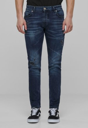 Jean slim fit bleu foncé avec détails déchirés, dégradé subtil, cinq poches et passants de ceinture, associés à des baskets noir et blanc.