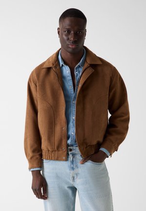 Homme portant une veste en daim marron sur une chemise en denim bleu clair déboutonnée et un jean bleu clair, debout devant un fond blanc uni.