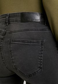 Jeans de ganga pretos com uma textura suave, apresentando costuras pronunciadas nos bolsos traseiros e um remendo de marca em couro na cintura.