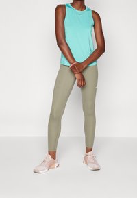 Na sobie znajduje się jasnoniebieski bezrękawnik, oliwkowe legginsy i różowe buty sportowe. Strój ma dopasowany krój i miękką teksturę.