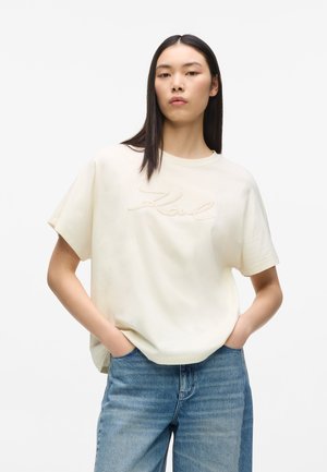 Ung kvinde med langt, glat hår iført en løs, cremefarvet T-shirt med Karl-logo og blå jeans, stående med hænderne i lommerne.