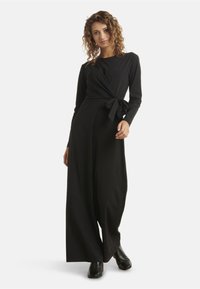 Combinaison noire avec des manches longues, design enveloppant, détail de lien à l'avant à la taille et pantalons à jambes larges, fabriquée en tissu lisse.