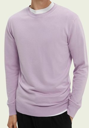 Mężczyzna w jasnofioletowym swetrze z okrągłym dekoltem na białej koszuli, w czarnych spodniach, na jednolitym jasnym tle.