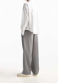 Een persoon die zijwaarts staat in een losse witte gestreepte shirt, brede grijze broek en witte sneakers tegen een witte achtergrond.