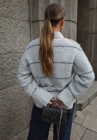 Hellgrauer, flauschiger Pullover mit dunklen Streifen, gerippte Bündchen, kombiniert mit dunklen Jeans und einer kleinen, gesteppten schwarzen Handtasche mit Kettenriemen.