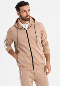 Homme avec barbe portant un sweat à capuche beige zippé et un pantalon assorti, regardant sur le côté, une main dans la poche et l'autre détendue.
