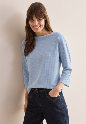 Haut bleu clair à côtes avec un col bateau et des manches trois-quarts, associé à un jean en denim foncé avec une coupe classique et des coutures visibles.