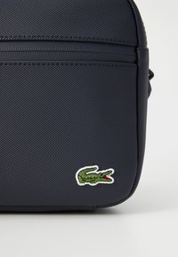 Geantă texturată de culoare albastru marin, cu un fermoar neted, având pe față un logo brodat cu un crocodil verde. Formă compactă și rectangulară.
