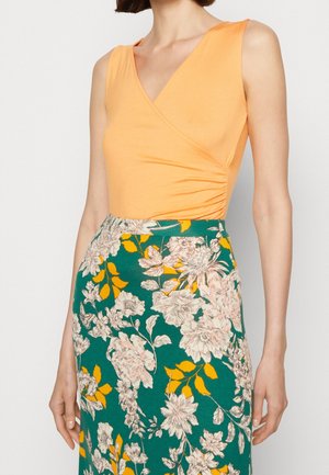 Frau trägt ärmelloses orangefarbenes Oberteil mit Wickeldetail und grünen Blumenrock mit gelben und weißen Blüten, Beschnitt an Hals und Handgelenken.