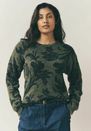 Green knit sweater featuring black floral patterns, round neckline, loose fit, and long sleeves, paired with blue denim pants and a belt.