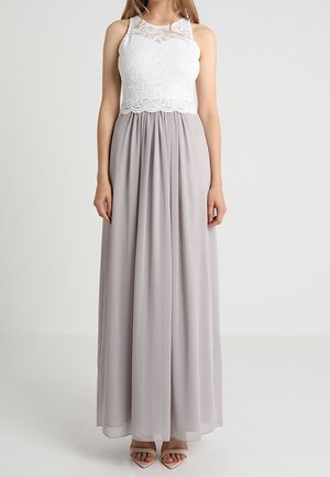 Kvinde iført hvid blonderbluse uden ærmer med høj hals og lang, flydende lysegrå nederdel, stående mod en ensfarvet baggrund.