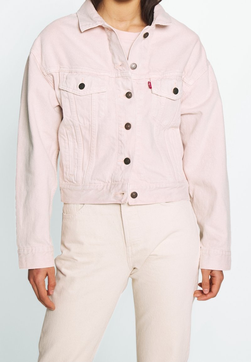 Femme portant une veste en jean rose clair boutonnée et un pantalon assorti de couleur claire, montrée du cou aux hanches.