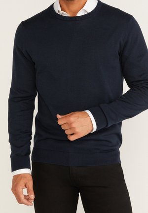 Hombre con suéter azul oscuro de cuello redondo sobre camisa blanca con cuello y pantalones negros, ajustando la manga del suéter cerca de la cintura.
