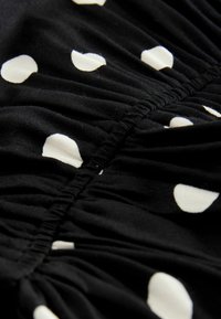 Tissu noir avec des pois blancs, présentant une texture plissée le long du centre, ajoutant de la dimension et des détails au design.