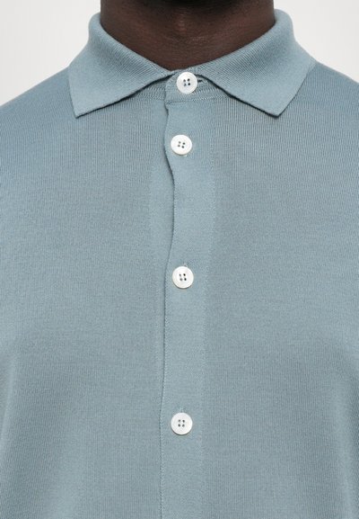 Polo em malha azul clara com gola, quatro botões brancos e tecido texturizado. Close-up mostra a pala dos botões da camisa.