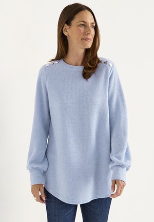 Lichtblauwe geribbelde trui met lange mouwen, ronde zoom en knoopdetails op de schouders. Draagt met donkerblauwe jeans.