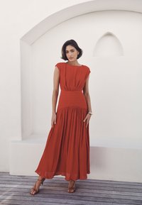 Červené maxi šaty s plisovaním a krátkymi rukávmi, priliehavo strihaný top a splývavá sukňa. Hladká textúra, dĺžka do polovice lýtok, v kombinácii so sandálmi na remienky.