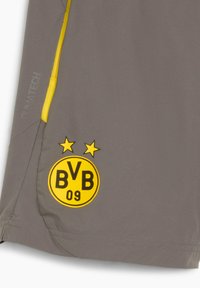 Grå shorts med en gul accentdragkedja, texturerad tyg och en svart/gul cirkulär logotyp med "BVB 09" och två stjärnor.