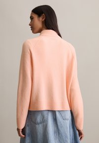 Jasnoróżowy sweter z wysokim kołnierzem i prążkowanym dołem, wykonany z miękkiego materiału, noszony z dżinsową spódnicą. Posiada rękawy raglanowe i luźny krój.