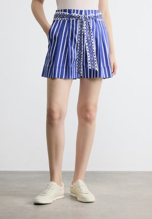 Blau-weiße Shorts mit vertikalen Streifen und einem gebundenen gepunkteten Gürtel, getragen mit weißen Sneakers und einem neutralen Oberteil, stehend auf einem grauen Boden.