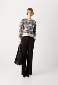 Maglione lavorato a maglia in crema con motivi geometrici neri, abbinato a pantaloni neri a vita alta. Modella che tiene una borsa nera texturizzata.