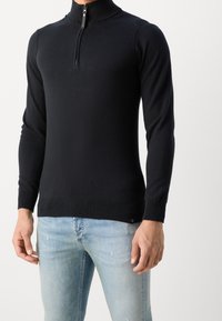 Schwarzer Pullover mit hohem Kragen und Viertelreißverschluss. Schmale Passform, gerippte Bündchen und Saum, aus einem glatten Stoff gefertigt. Kombiniert mit hellblauen Jeans.