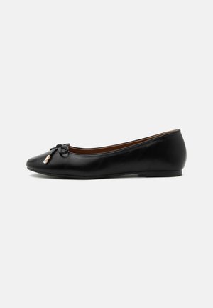 Ballerine plate en cuir noir avec un bout arrondi, un petit nœud aux extrémités dorées et un talon plat et bas sur fond gris.