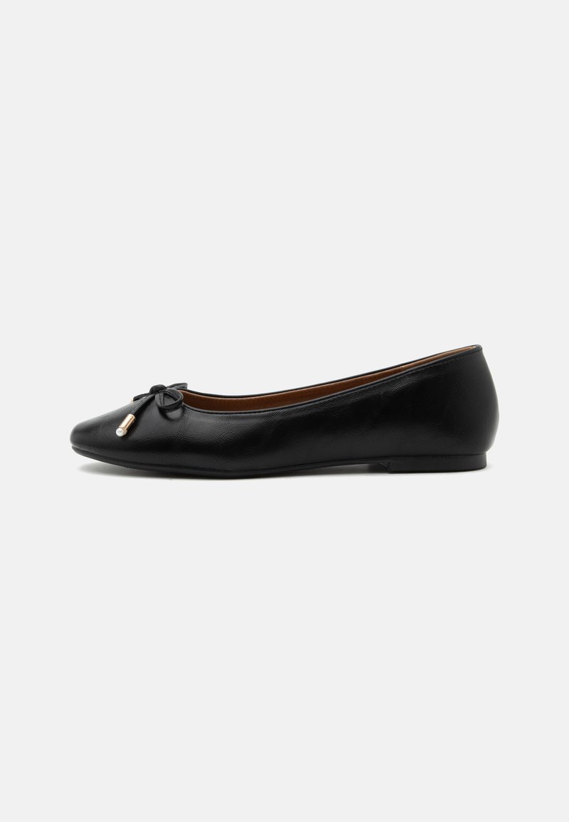 Ballerine plate en cuir noir avec un bout arrondi, un petit nœud aux extrémités dorées et un talon plat et bas sur fond gris.