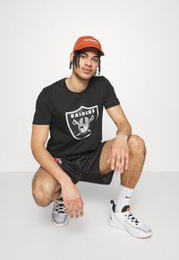 Camiseta negra con un logo de los Raiders en blanco y gris, combinada con pantalones cortos negros y zapatillas multicolores. Gorra de color óxido.